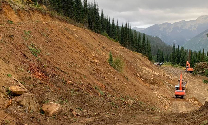 Digging-on-Hillside a large sand and dirt pile with coniferous trees at the base and mountains seen in the distance. also at the base of the pile are 2 large excavators.